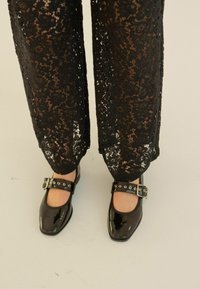 Černé lakované kožené baleríny typu Mary Jane se zaoblenou špičkou a širokým kotníkovým páskem s stříbrnými detaily, doplněné černými krajkovými kalhotami.