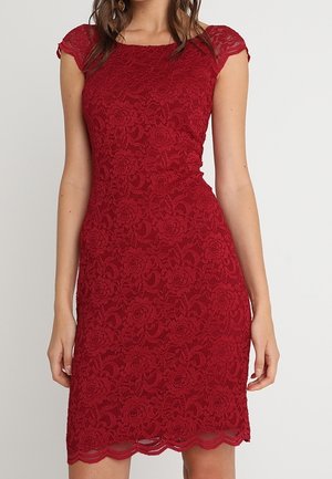 Dopasowana, czerwona sukienka z koronki, sięgająca do kolan, z krótkimi rękawkami typu kaptur i karbowanym dołem, noszona przez kobietę o falowanych brązowych włosach.