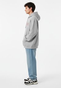 Felpa grigia con cappuccio e testo rosso, tasca a marsupio e vestibilità ampia, abbinata a jeans azzurri e scarpe da ginnastica bianco e nere.