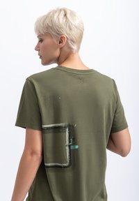 T-shirt en coton vert olive avec un imprimé graphique d'un motif de tuyau en gris et turquoise au dos. Manches courtes et col rond.