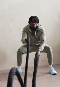 Gray athletic tracksuit, textured fabric, lightweight design. Black battle ropes held in hands, light blue sneakers. Neutral background.