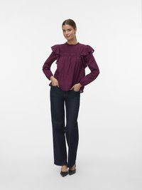 Blouse bordeaux à volants avec un motif texturé, des détails plissés et un col rond, associée à un pantalon en denim foncé évasé et des escarpins noirs.