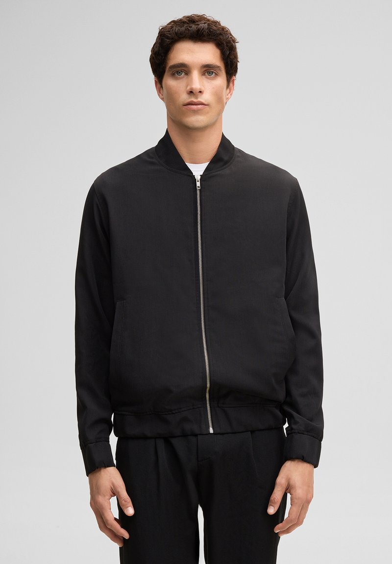 Junger Mann mit lockigem Haar, der eine schwarze Bomberjacke mit Reißverschluss und schwarze Hosen trägt, vor einem schlichten grauen Hintergrund steht.