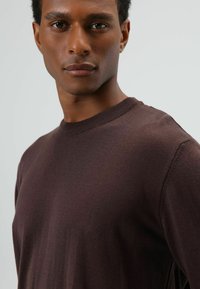 Pull marron foncé avec un col rond, un col côtelé et une texture lisse, présenté en gros plan sur le haut du corps d'un modèle sur un fond clair.