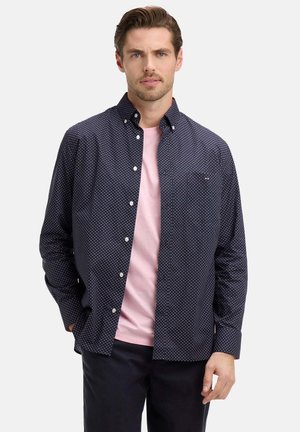 Homme portant une chemise bleue marine à pois boutonnée sur un t-shirt rose clair et un pantalon foncé sur un fond blanc uni.