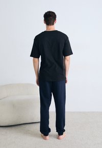 Schwarzes Baumwoll-T-Shirt mit Rundhalsausschnitt, kombiniert mit marineblauen Jogginghosen. Das Model steht barfuß, mit dem Rücken zur Kamera, in einer hellen Innenumgebung.