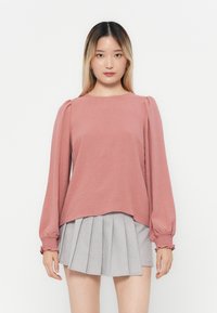 Rosa, strukturert langermet topp med pufferte skuldre, kombinert med et grått plissert skjørt. Toppen har rund hals og elastiske mansjetter.