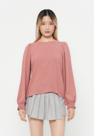 Rosa, strukturiertes Langarmoberteil mit puffigen Schultern, kombiniert mit einem grauen, plissierten Rock. Das Oberteil hat einen runden Halsausschnitt und elastische Bündchen.