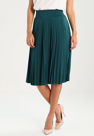 Grønblå plissert midi-nederdel båret med hvid top, vist med korslagte ben i nøgenfarvede højhælede sko mod en ensfarvet baggrund.