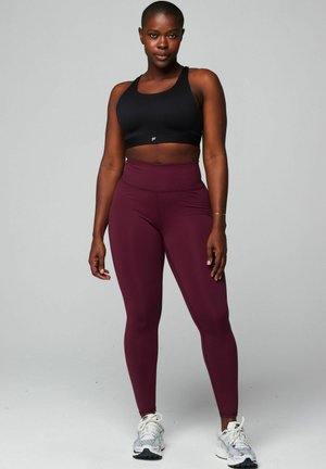 Schwarzer Sport-BH mit breitem Bund und Mesh-Details, kombiniert mit hoch taillierten bordeauxroten Leggings, und dazu weiße Sportschuhe.