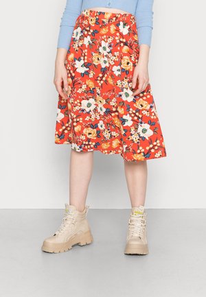 Jupe midi fleurie rouge avec des fleurs jaunes et bleues, taille élastique, associée à des bottines chunky beiges. Tissu lisse, longueur genou.