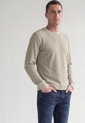 Hombre con camisa beige de manga larga y vaqueros azules, de pie con las manos en los bolsillos frente a un fondo claro y liso.