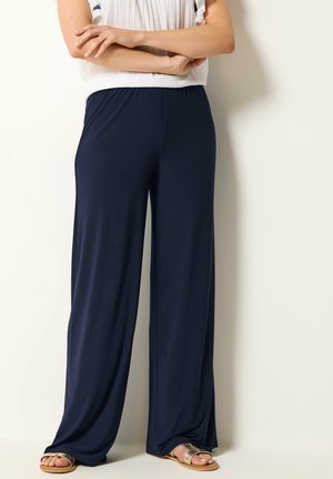 Femme debout, bras croisés, portant un haut blanc sans manches, un pantalon large bleu marine et des sandales beiges sur un fond clair uni.