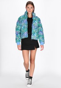 Giacca a vento accorciata in stampa leopardata multicolore con dettagli blu, caratterizzata da un colletto alto e chiusura a zip. La modella indossa un abito nero.