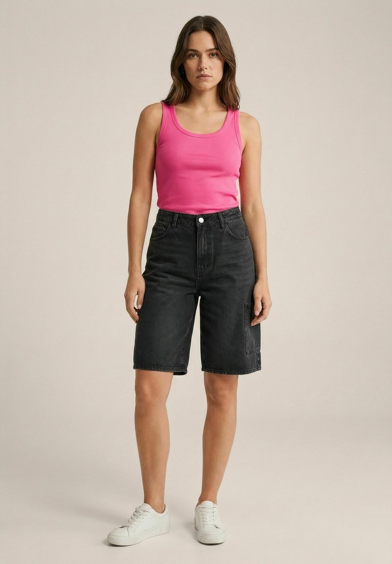 Femme debout, de face, vêtue d'un haut rose sans manches, d'un short en jean noir au genou et de baskets blanches sur un fond uni.