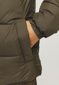 Veste matelassée vert olive avec une texture douce, dotée d'une poche latérale et de poignets élastiqués pour la chaleur et le confort.