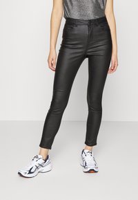 Person som bär svarta tajta läderbyxor och vita löparskor med marinblå och orange inslag, står mot en enkel bakgrund.