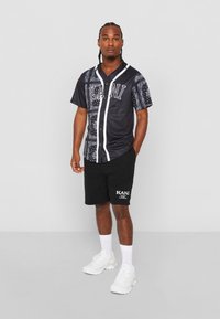 Sort kortærmet skjorte med knaplukning og hvid kant samt bandana-mønstrede detaljer, parret med sorte shorts med et logo. Hvide sneakers.