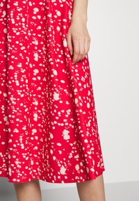 La mano de una mujer reposando sobre una falda roja con manchas abstractas blancas, contra un fondo blanco liso.