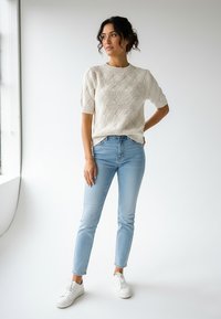 Kremowy sweter dzianinowy z fakturowanym wzorem, krótkie rękawy, w połączeniu z jasnoniebieskimi dżinsami i białymi trampkami. Model stoi na tle jednolitego tła.