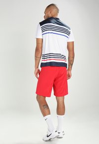Pánsky športový outfit zložený z bieleho polo trička s námorníckymi a červenými horizontálnymi pruhmi, doplnený červenými šortkami, bielymi ponožkami a čiernymi teniskami.