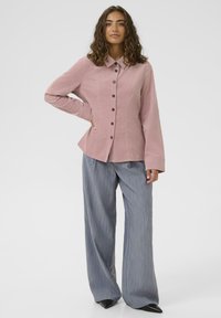 Žena s kovrčavom kosom nosi rozu košulju na dugmad i široke sive hlače s prugama, stoji s jednom rukom na boku.