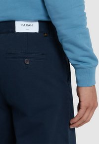 Navy broek met een achterzak, voorzien van een bruine knop en een wit label met de tekst "FARAH." De drager heeft een lichtblauwe trui aan.
