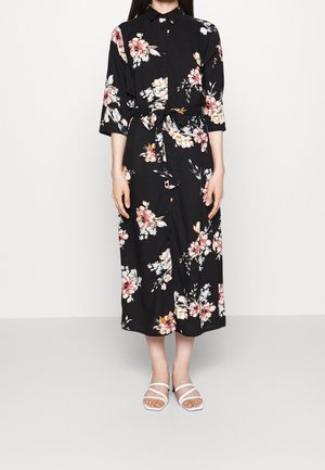 Zwarte midi overhemdjurk met roze en witte bloemenprint, driekwart mouwen, tailleceintuur, gedragen met witte sandalen met bandjes.