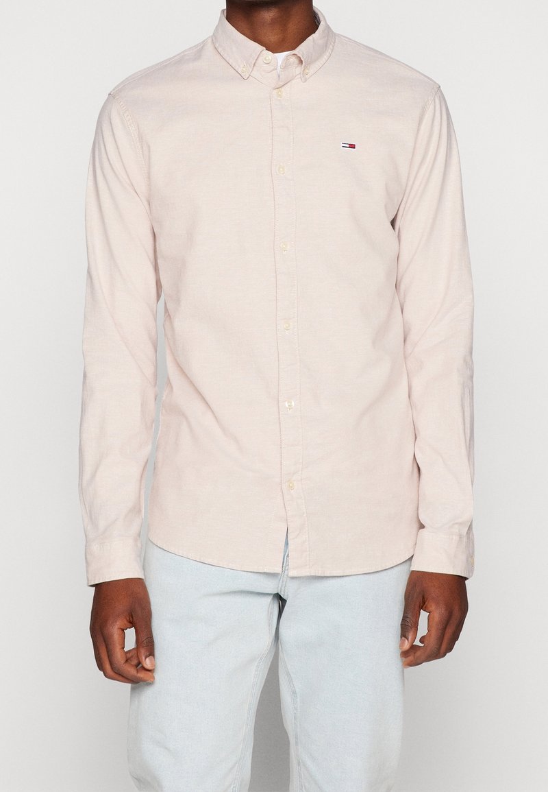 Homme portant une chemise à manches longues beige clair avec un petit logo sur la poitrine et un jean bleu clair, debout devant un fond uni.