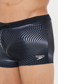 Schwarze Bade shorts mit einem Wirbelmuster in Grautönen. Hergestellt aus glattem Material mit einem Kordelzugbund und einem weißen Speedo-Logo.