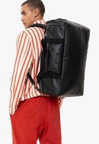 Czarny materiałowy plecak o prostokątnym kształcie, z zasuwanymi kieszeniami i mocnymi paskami. Ma naszywkę z logo z boku. Gładka faktura.