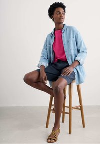 Denim button-up shirt layered over a pink top, paired with dark shorts and yellow strappy sandals, seated on a wooden stool.