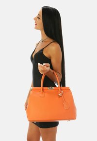 Oranssi nahkainen käsilaukku, jolla on jämäkkä muoto, kultainen varustus ja kaksoiskahvat. Pintakäsittely on sileä ja siinä on hienovaraisia yksityiskohtia ylhäällä.