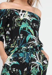 Kvinna i en svart avknäppt topp och byxor med grön- och vitblommigt mönster, med händerna avslappnat i fickorna.