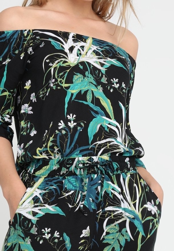 Kvinna i en svart avknäppt topp och byxor med grön- och vitblommigt mönster, med händerna avslappnat i fickorna.