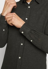 Camicia grigia scuro con bottoni, realizzata in tessuto strutturato, caratterizzata da un colletto classico e bottoni bianchi sui polsini e sulla patta.