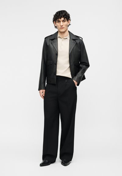 Νεαρός άνδρας με σγουρά μαλλιά που φοράει μαύρο δερμάτινο μπουφάν, μπεζ πόλο πουκάμισο, φαρδιά μαύρη παντελόνια και μαύρα παπούτσια, στέκεται με το ένα χέρι στην τσέπη.