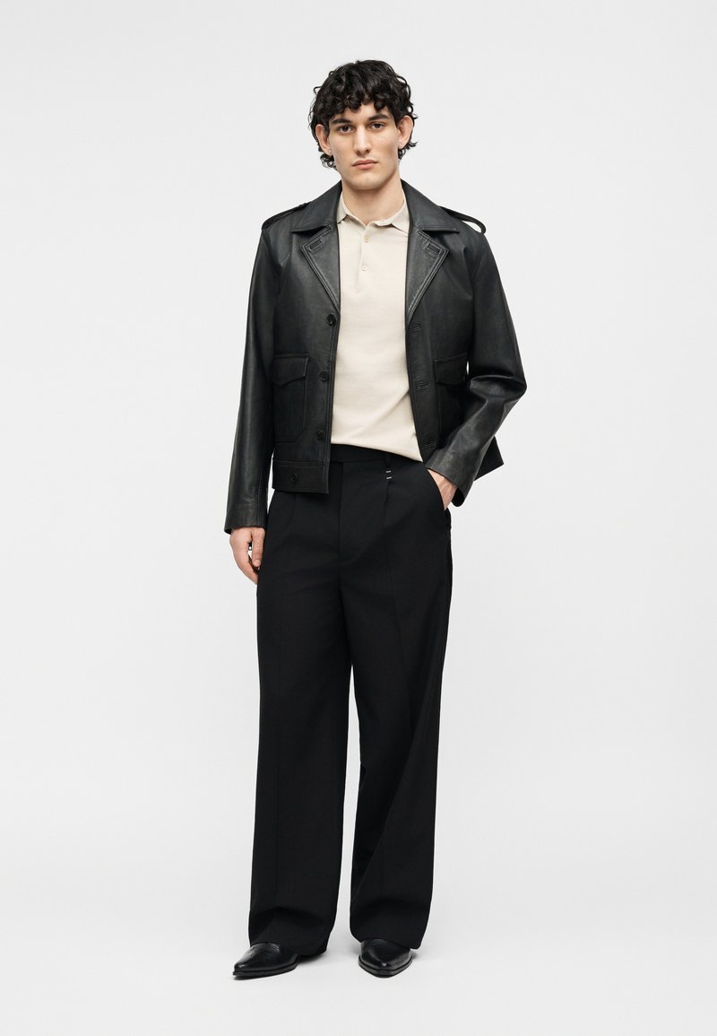 Young man with curly hair wearing black leather jacket, beige polo shirt, wide black trousers, and black shoes, standing with one hand in pocket.