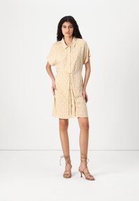 Kortermet, krageklær i lys beige med skjellmønster og innsnevret midje. Kombinert med strappy beige hæler.