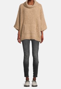 Beżowy, strukturalny, oversize'owy sweter z kapturem, z szerokimi rękawami 3/4 i kieszeniami z przodu, zestawiony z wąskimi szarymi dżinsami i jasnymi butami.