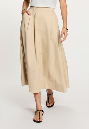 Falda midi beige con pequeños detalles bordados, combinada con top blanco y sandalias negras de tiras en T, complementada con pulseras doradas.