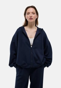 Sweat à capuche zippé bleu marine avec une coupe oversize, capuche à cordon de serrage et texture douce, associé à un débardeur blanc en dessous.