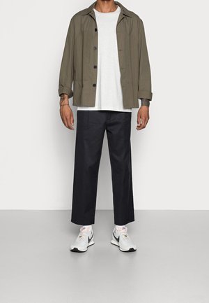 Mann in olivgrünem, knopfbarem Sakko, weißem T-Shirt, schwarzen weit geschnittenen Hosen, weißen Nike Turnschuhen und weißen Socken steht vor einer weißen Wand.