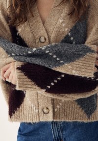 Isik, kes kannab beežikat kardigani, millel on suured nööbid ja tumeda halli ning burgundia argyle-muster, käed risti rinnal.