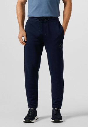 Hombre con pantalones de chándal azul marino con cordón y bolsillos laterales, y zapatillas negras, de pie frente a un fondo claro y liso.