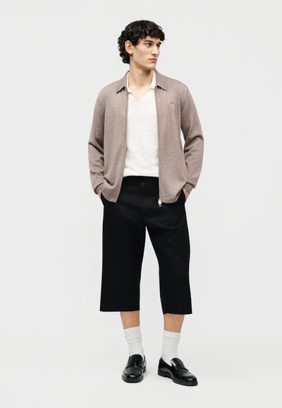 Νέος με σγουρά μαλλιά που φοράει μπεζ ζακέτα με φερμουάρ, λευκό πουκάμισο, μαύρο φαρδύ παντελόνι 3/4, λευκές κάλτσες και μαύρα μοκασίνια, όρθιος με τα χέρια στις τσέπες.