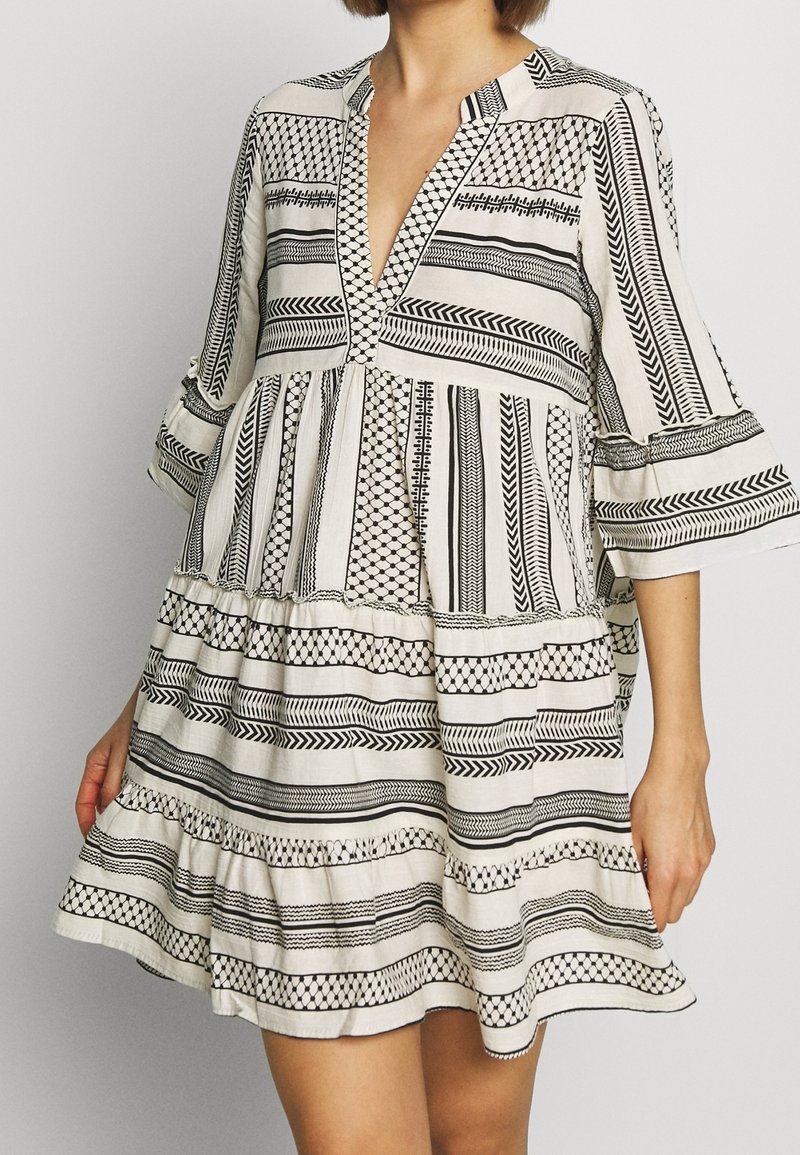 Femme portant une robe blanche large avec des motifs géométriques et rayés noirs, des manches trois-quarts évasées et un décolleté en V profond.