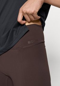 Brune leggings med en jevn tekstur og en midjebånd med et subtilt preget logo. En løs mørk topp er lett hevet for å avsløre leggingsene.