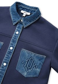 Marineblaue Hemd mit einem Denimkragen und einer Tasche. Die Tasche hat ein geprägtes Design. Die vorderen Knöpfe sind weiß und heben sich vom Stoff ab.