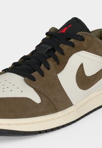 Χαμηλό sneaker με καφέ επενδύσεις από σουέτ, λευκά δερμάτινα πάνελ, μαύρα κορδόνια, κόκκινο λογότυπο Jumpman στη μαύρη γλώσσα και εκρού σόλα με μαύρη εξωτερική σόλα.